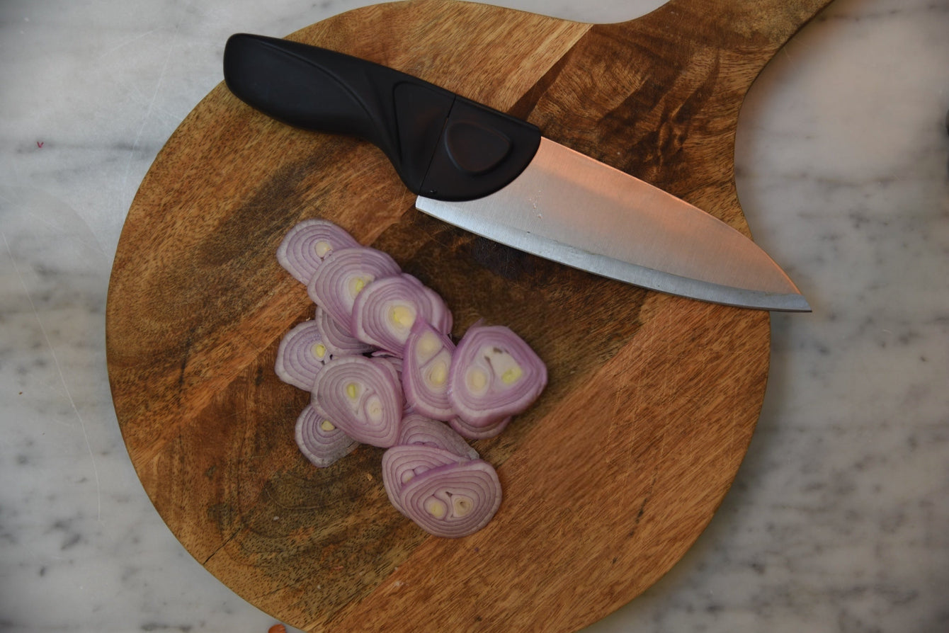 A Gray Chef knife on a Round cutting board with thin shallots on a marble counter