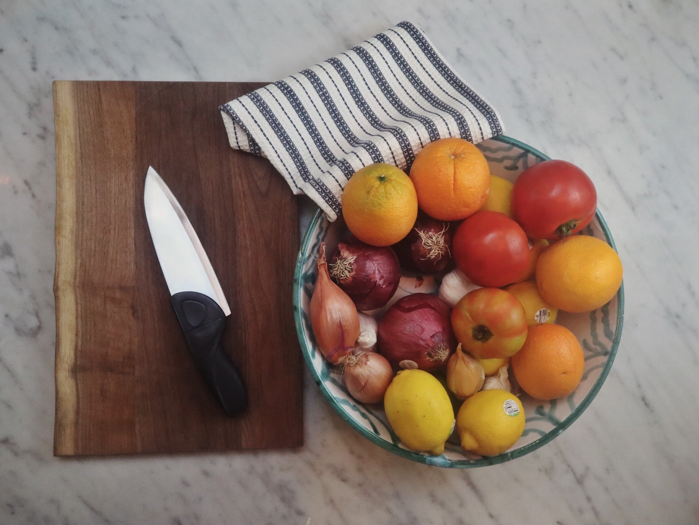 A Grey Chef Knife with a Waffle Striped Navy Kitchen Towel, a Bowl of mixed onion, shallot, tomatoes and citrus, a chef knife for women, on a wood cutting board on a marble countertop