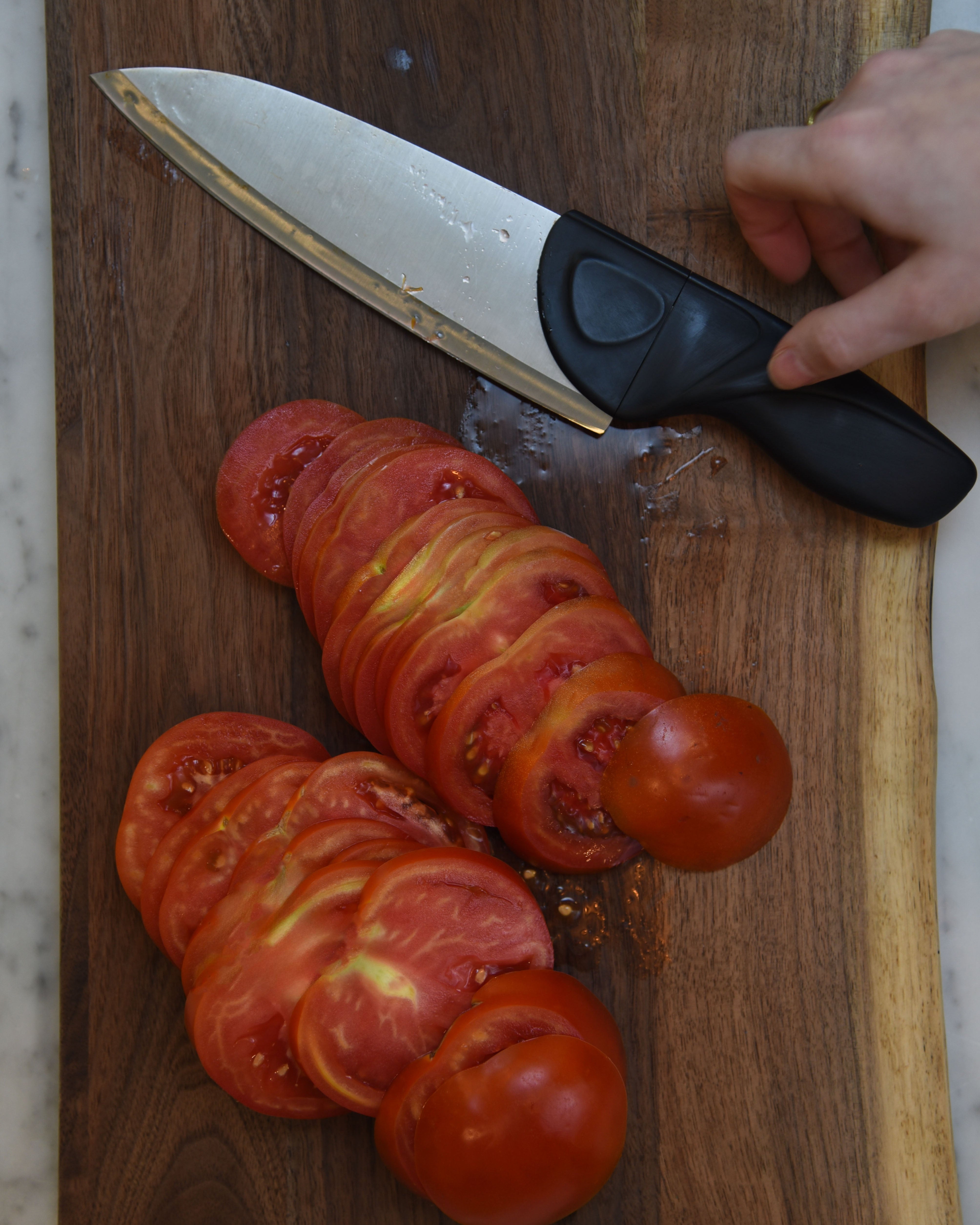 Sliced tomatoes on a wooden cutting board with a knife and a hand holding it.