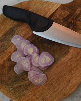A Gray Chef knife on a Round cutting board with thin shallots on a marble counter