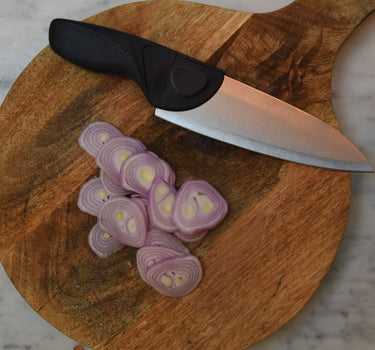 A Gray Chef knife on a Round cutting board with thin shallots on a marble counter