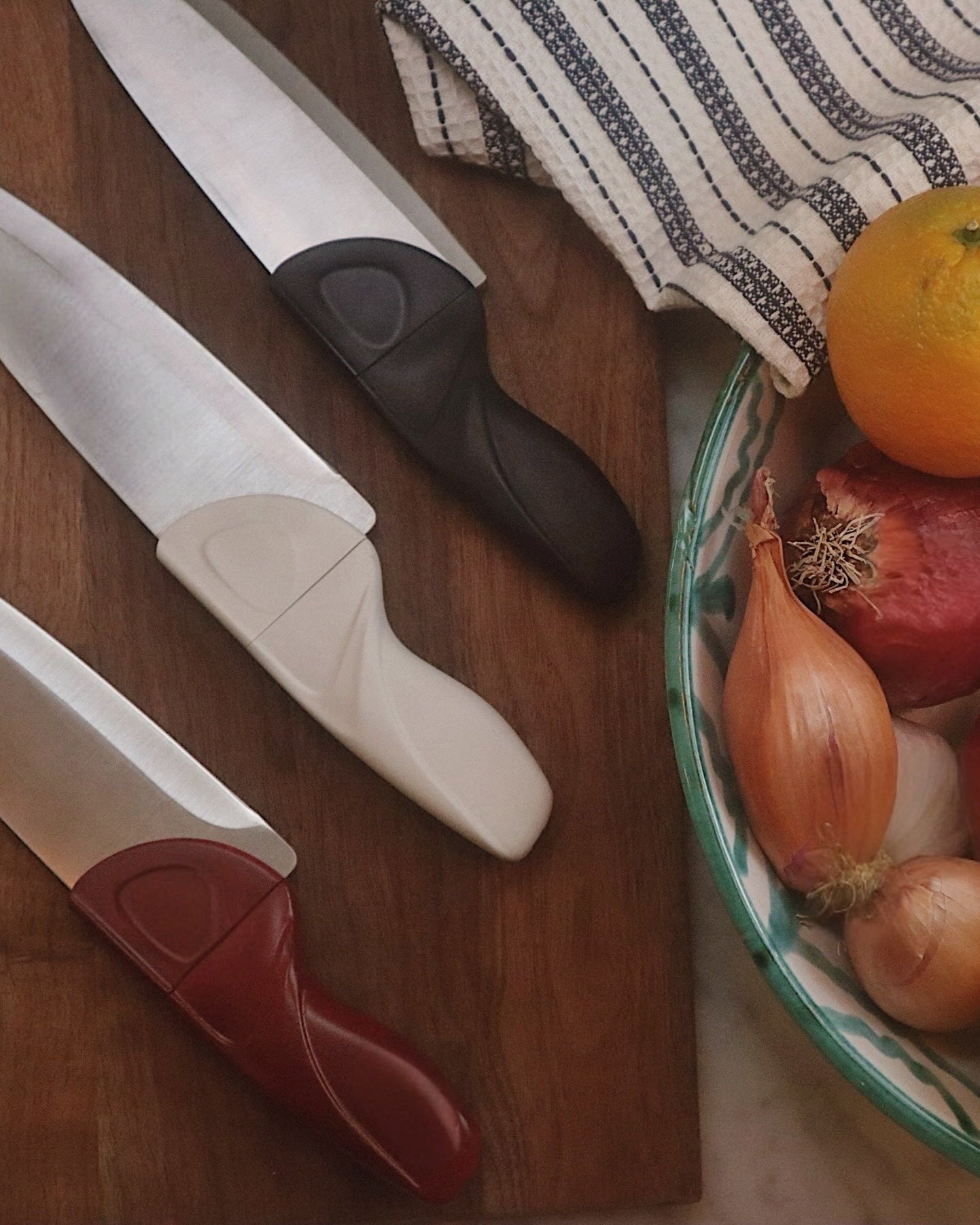Fruits and vegetables in a bowl with kitchen knives and a cutting board on a wooden surface.