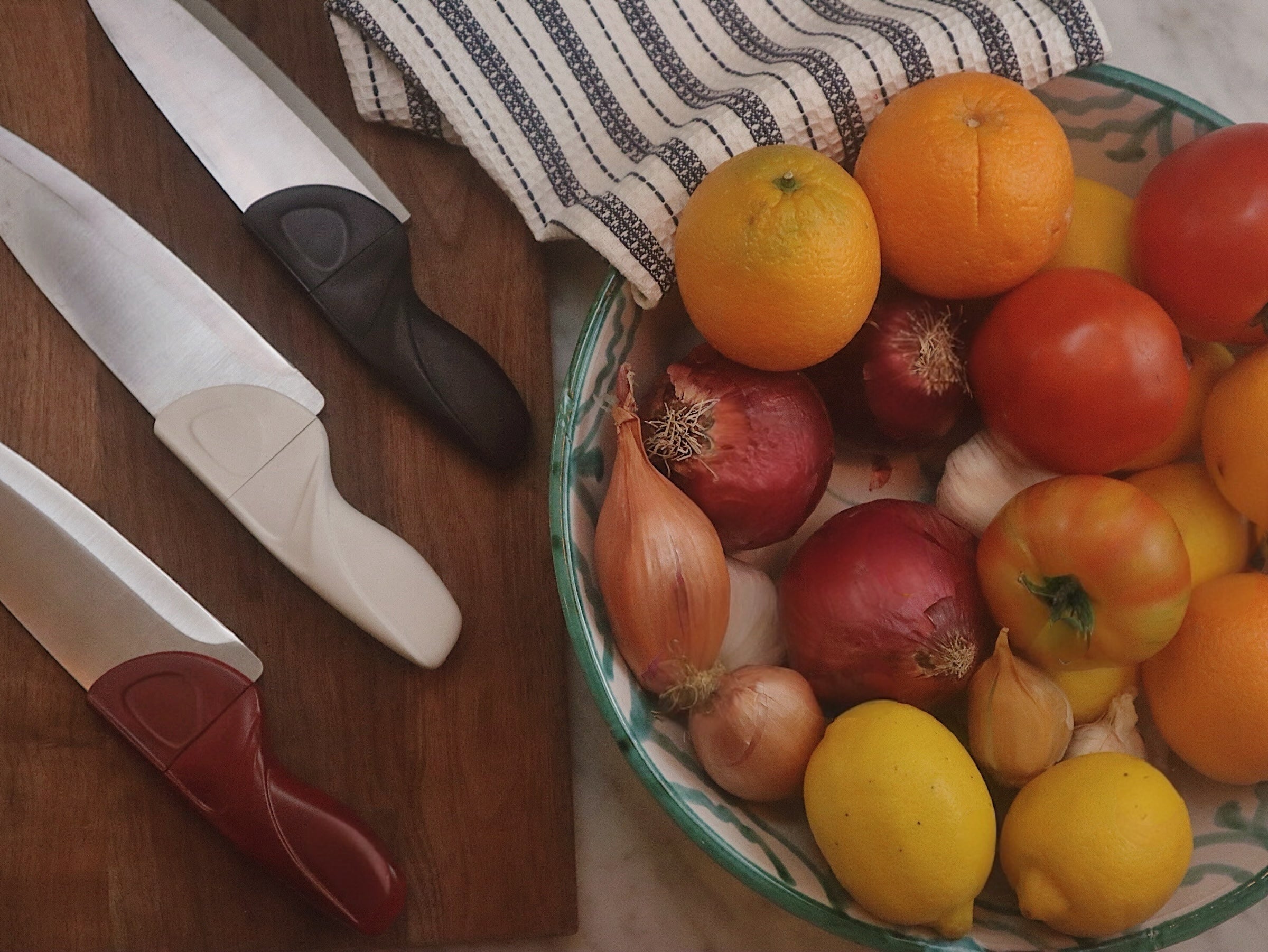 Fruits and vegetables in a bowl with kitchen knives and a cutting board on a wooden surface.