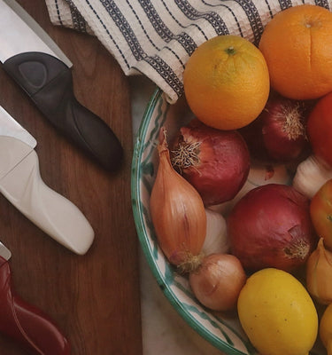 Fruits and vegetables in a bowl with kitchen knives and a cutting board on a wooden surface.