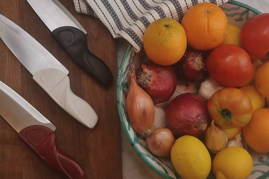 Fruits and vegetables in a bowl with kitchen knives and a cutting board on a wooden surface.