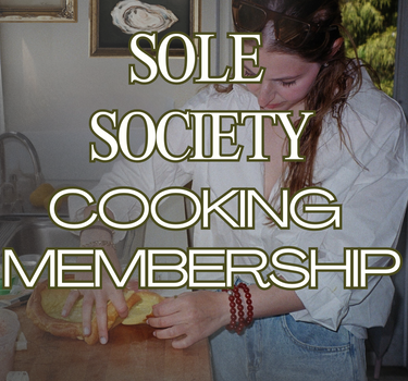 A young woman prepares a rustic golden-brown Dutch baby pancake on a butcher block counter, surrounded by a lived-in, sunlit kitchen. She wears a relaxed white blouse and denim jeans, with tortoiseshell sunglasses tucked into her hair and red beaded bracelets on her wrist. Framed still-life paintings of a lemon, oysters, and sardines hang on the wall behind her, adding to the nostalgic, European-inspired kitchen aesthetic. Sunlight streams in from a window, highlighting the greenery outside. Overlaid text r
