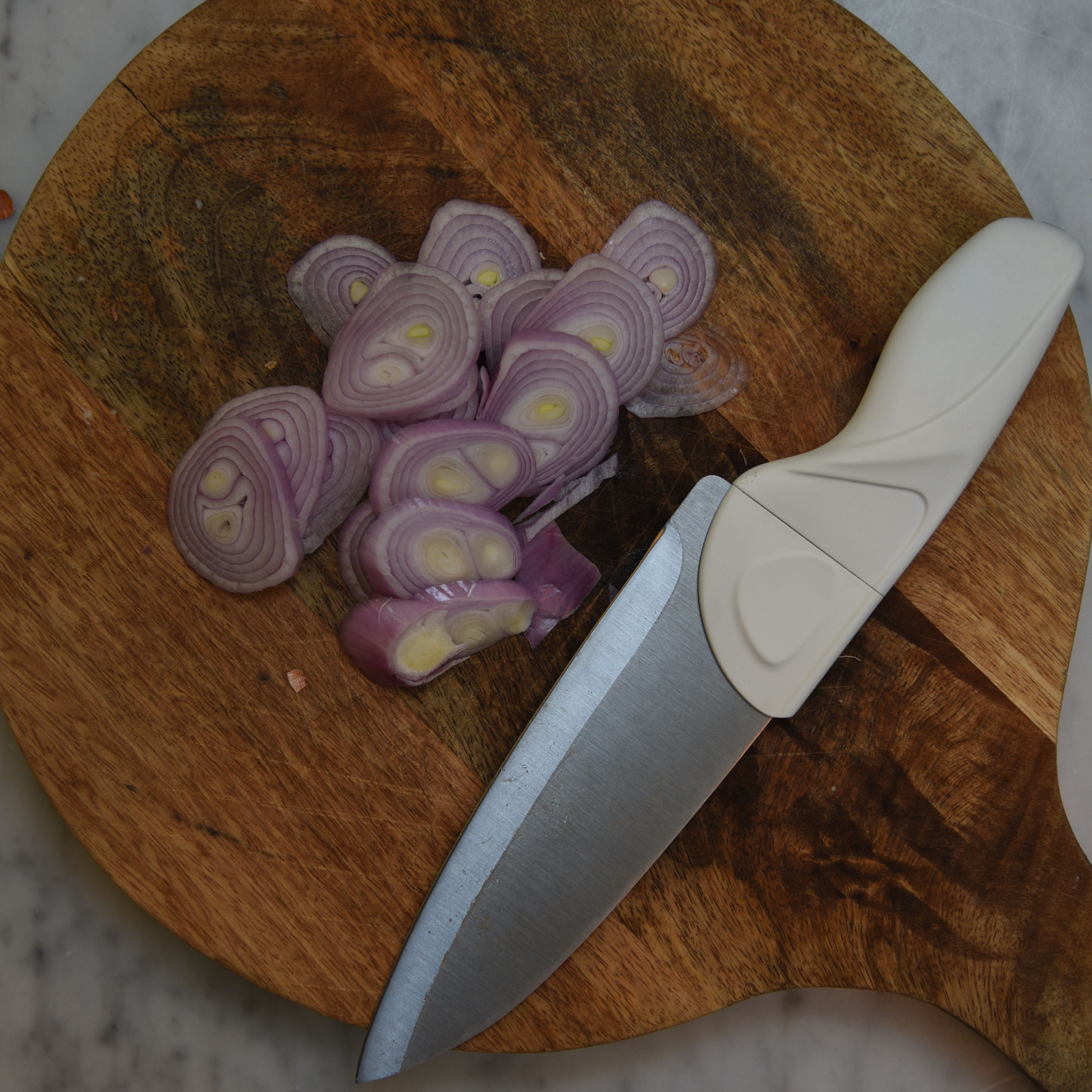 Cream Handle Chef Knife cut shallots thin sliced, on round wood cutting board with handle on marble countertop