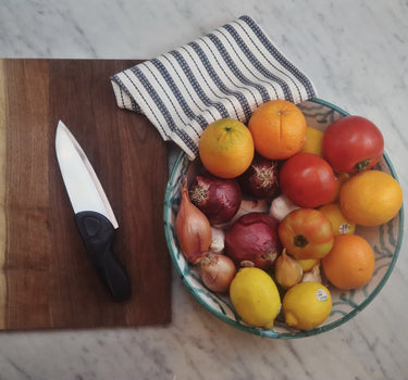 A Grey Chef Knife with a Waffle Striped Navy Kitchen Towel, a Bowl of mixed onion, shallot, tomatoes and citrus, a chef knife for women, on a wood cutting board on a marble countertop