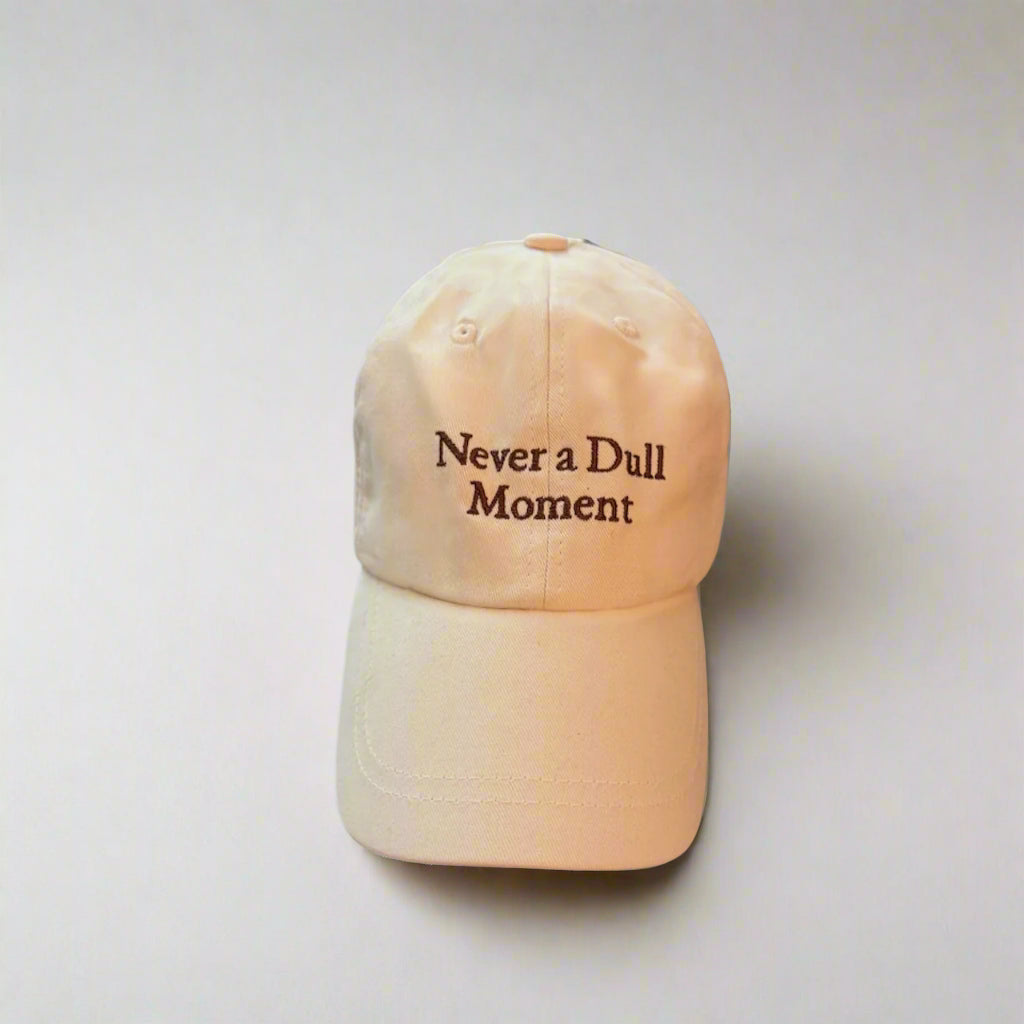 Beige cap with 'Never a Dull Moment' text on a light gray background