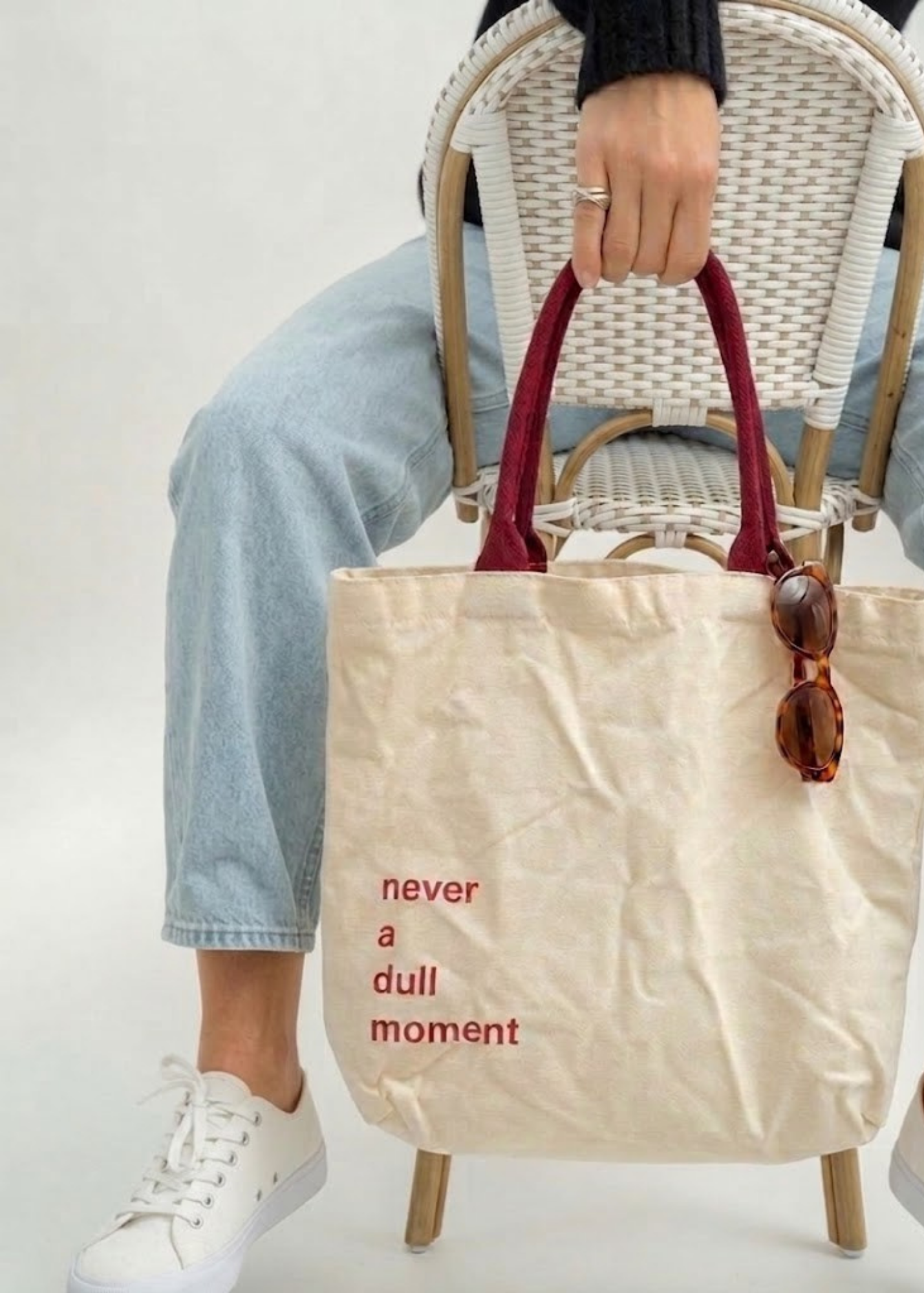 Person sitting on a wicker chair holding a beige tote bag with red handles.