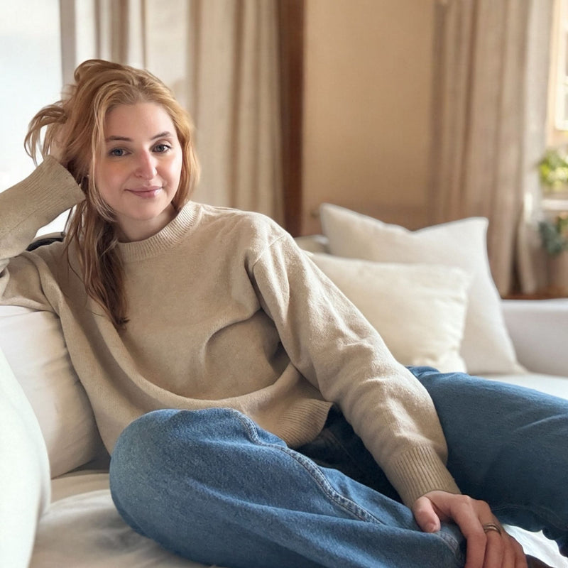 Woman sitting on a couch wearing a beige sweater and blue jeans, with a neutral-colored room in the background.
